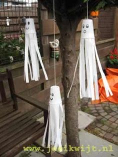 three white paper ghost decorations hanging from a tree in front of a bench and flower pot