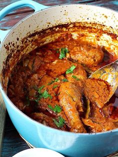 a blue pot filled with meat and sauce on top of a wooden table next to spoons