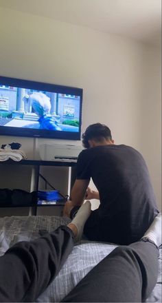 a man sitting in front of a flat screen tv with his feet propped up on the bed