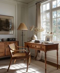 a wooden desk sitting in front of a window next to a chair and table with a lamp on it