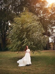 a woman in a white dress is standing in the grass with trees and bushes behind her