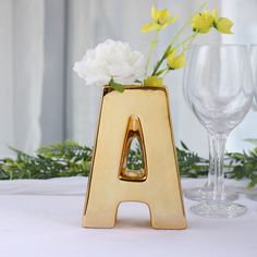 a gold letter shaped vase with flowers in it and two wine glasses on the table