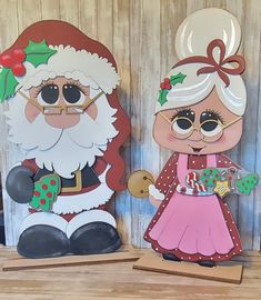 two paper cutouts of santa and mrs claus on wooden planks with christmas decorations