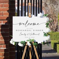 a welcome sign with white flowers on it in front of a brick wall and gate