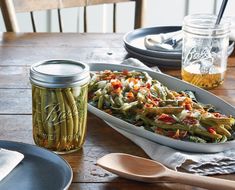 a wooden table topped with plates and bowls filled with food next to a jar of pickled beans