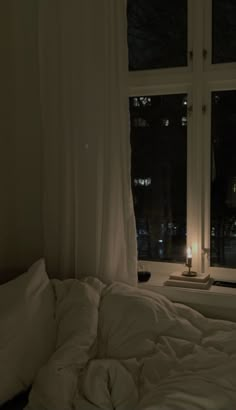 an unmade bed sitting in front of a window next to a night stand with a lit candle