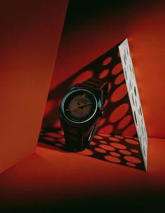 a red watch sitting on top of an orange surface next to a white box with a shadow