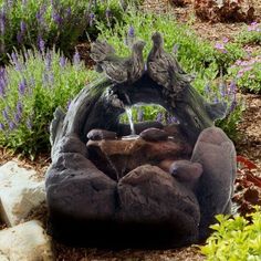 a water fountain in the middle of a garden with rocks and plants around it, surrounded by purple flowers