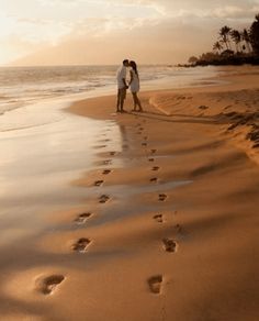 a man and woman standing on top of a beach next to the ocean with footprints in the sand