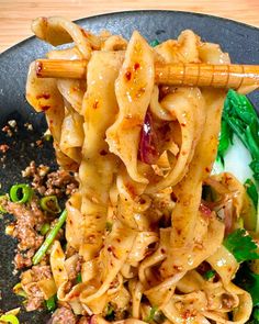 noodles with meat and vegetables in a black bowl on top of a wooden table next to chopsticks