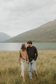 a man and woman standing next to each other in tall grass near water with mountains in the background