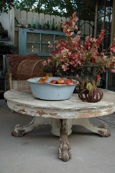 a bowl of fruit is sitting on an old table with wicker chairs and flowers in the background