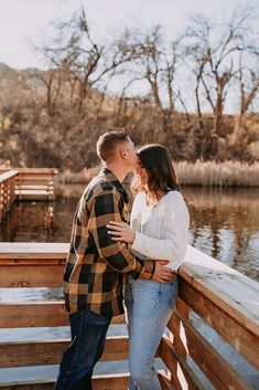 a man and woman standing next to each other on a wooden bridge near water with trees in the background