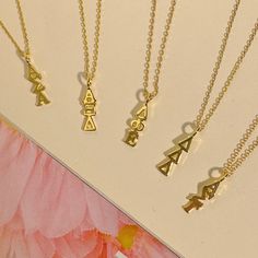 four gold necklaces with letters on them sitting next to a pink and white flower