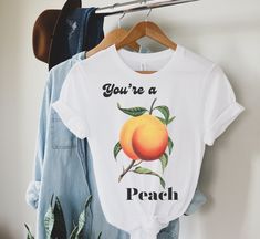 Introducing our delightful t-shirt that says You're a peach with a charming peach graphic! This adorable t-shirt is the perfect way to spread some positivity and sweet vibes wherever you go. Made from soft, high-quality fabric, this shirt will keep you comfortable all day long. Whether you're grabbing brunch with friends or simply lounging at home, this t-shirt is a fun and stylish addition to any outfit. Show off your love for juicy peaches and let everyone know how peachy-keen you are with this fabulous tee. Perfect as a gift for your bestie or as a treat for yourself, this t-shirt is a must-have addition to any peach lover's wardrobe! .: Made using 100% US cotton that is ethically grown and harvested. Gildan is also a proud member of the US Cotton Trust Protocol ensuring ethical and sus Trendy Peach T-shirt With Letter Print, Trendy Peach Relaxed Fit T-shirt, Trendy Relaxed Fit Peach T-shirt, Peach Letter Print T-shirt For Spring, Summer Peach Cotton Shirt, Peach Cotton Summer Shirt, Cute Peach T-shirt With Graphic Print, Trendy Peach Tops With Letter Print, Trendy Peach Top With Letter Print