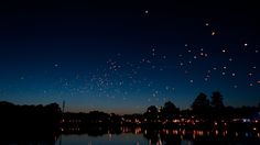 many balloons are flying in the sky above water at night with lights reflecting on the surface