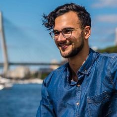 a man wearing glasses is sitting by the water with a bridge in the back ground