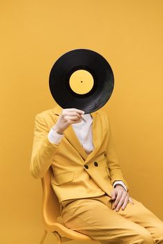 a man in a yellow suit and black hat sitting on a chair with his hand up to his face