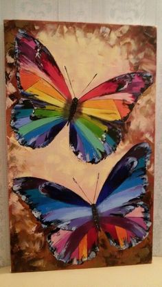 two colorful butterflies sitting on top of a wooden plaque in front of a white wall