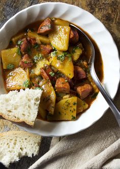 a white bowl filled with stew and bread on top of a table next to a spoon