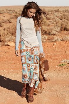 a woman walking across a dirt road in the middle of the desert with her hair blowing in the wind