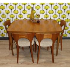 a wooden table with four chairs and a vase on it's end, in front of a wallpapered background