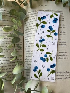 a bookmark with blue flowers and green leaves on top of an open book next to eucalyptus