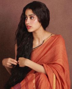 a woman with long black hair wearing an orange sari and holding her hand on her shoulder