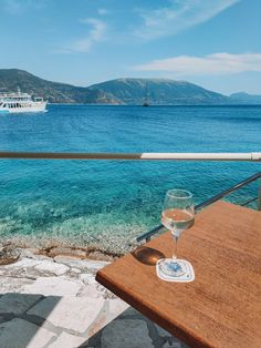 a glass of wine sitting on top of a wooden table next to the ocean with a boat in the background