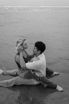 a man and woman sitting on the beach next to each other, looking at each other