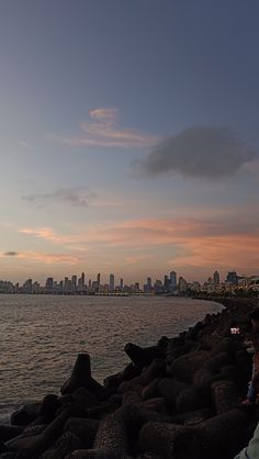 two people sitting on rocks near the water at sunset with city in the back ground