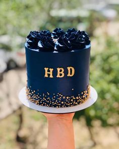 a hand holding up a blue and gold cake with the word hbd on it