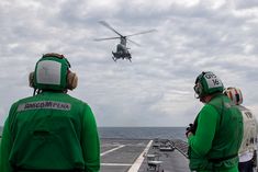 two men in green jackets watch as a helicopter flies over an aircraft carrier