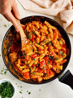 a skillet filled with pasta and vegetables being stirred by a wooden spoon on a white surface