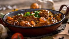 a bowl filled with meat and vegetables on top of a wooden table next to tomatoes