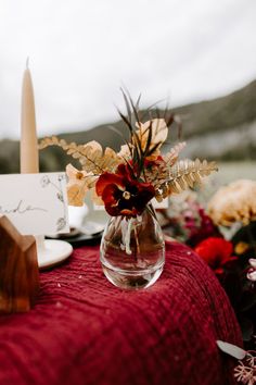 a vase filled with flowers sitting on top of a red table cloth next to candles