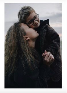 a woman with long hair and glasses is kissing another woman's face in front of an overcast sky