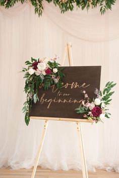 an easel with flowers and greenery on it in front of a white backdrop