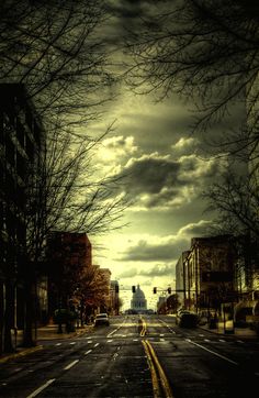 an empty street with no cars on it and dark clouds in the sky over buildings