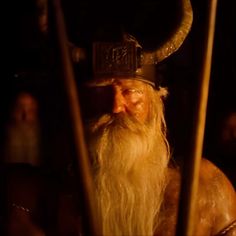 an old man with long white hair wearing a horned hat and holding two wooden poles