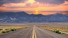 the sun is setting on an empty road in the desert with mountains in the background