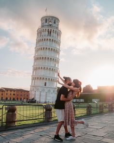 two people hugging each other in front of a leaning tower with the sun setting behind them