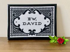 a cross stitch picture frame with a single red rose in front of it on a wooden table