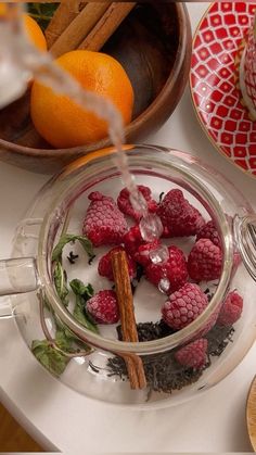 a glass teapot filled with raspberries next to oranges and cinnamon sticks