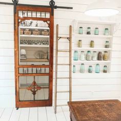 An ornately-carved sliding farmhouse pantry door with large glass panels is set into a white shiplap wall. Shelving with organized pulses and grains is set to the right of the pantry. A ladder leans against the wall...   Image: farmhouse2020