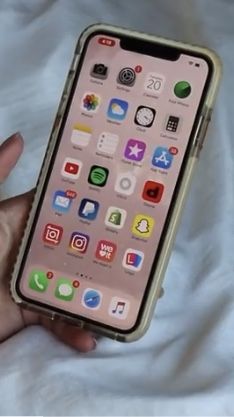 a person holding an iphone in their hand on a white sheet covered bed with the screen showing icons