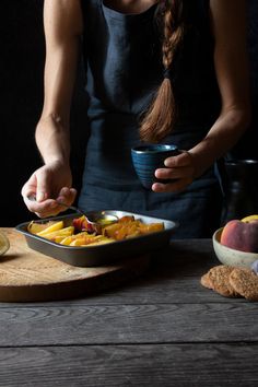 a woman is holding a spoon over a bowl of food on a wooden table with fruit