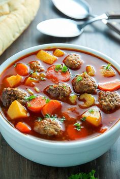 a white bowl filled with meatballs and vegetables next to bread on a wooden table