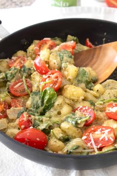 a pan filled with pasta and vegetables on top of a white table cloth next to a wooden spoon