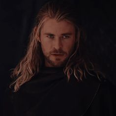 a man with long hair wearing a black cloak and looking at the camera in a dark room